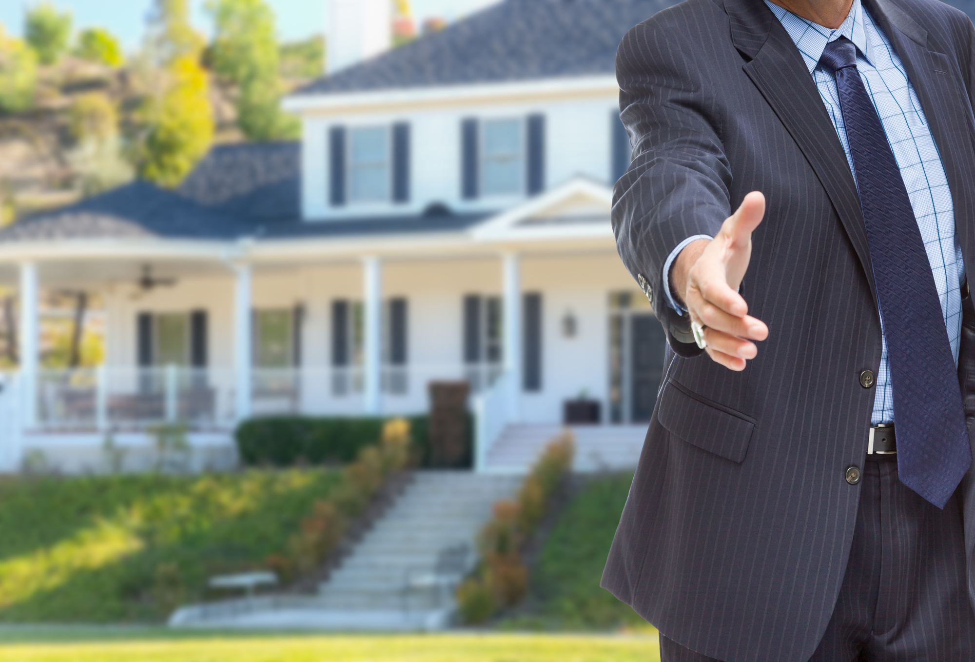 A realtor reaching toward the camera for a handshake with a beautiful house in the background.