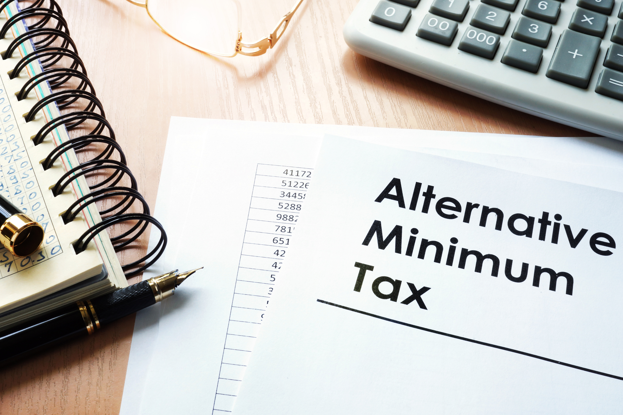 A document titled “alternative minimum tax” sits on a desk next to a spreadsheet of financial figures, a calculator, and a notebook.