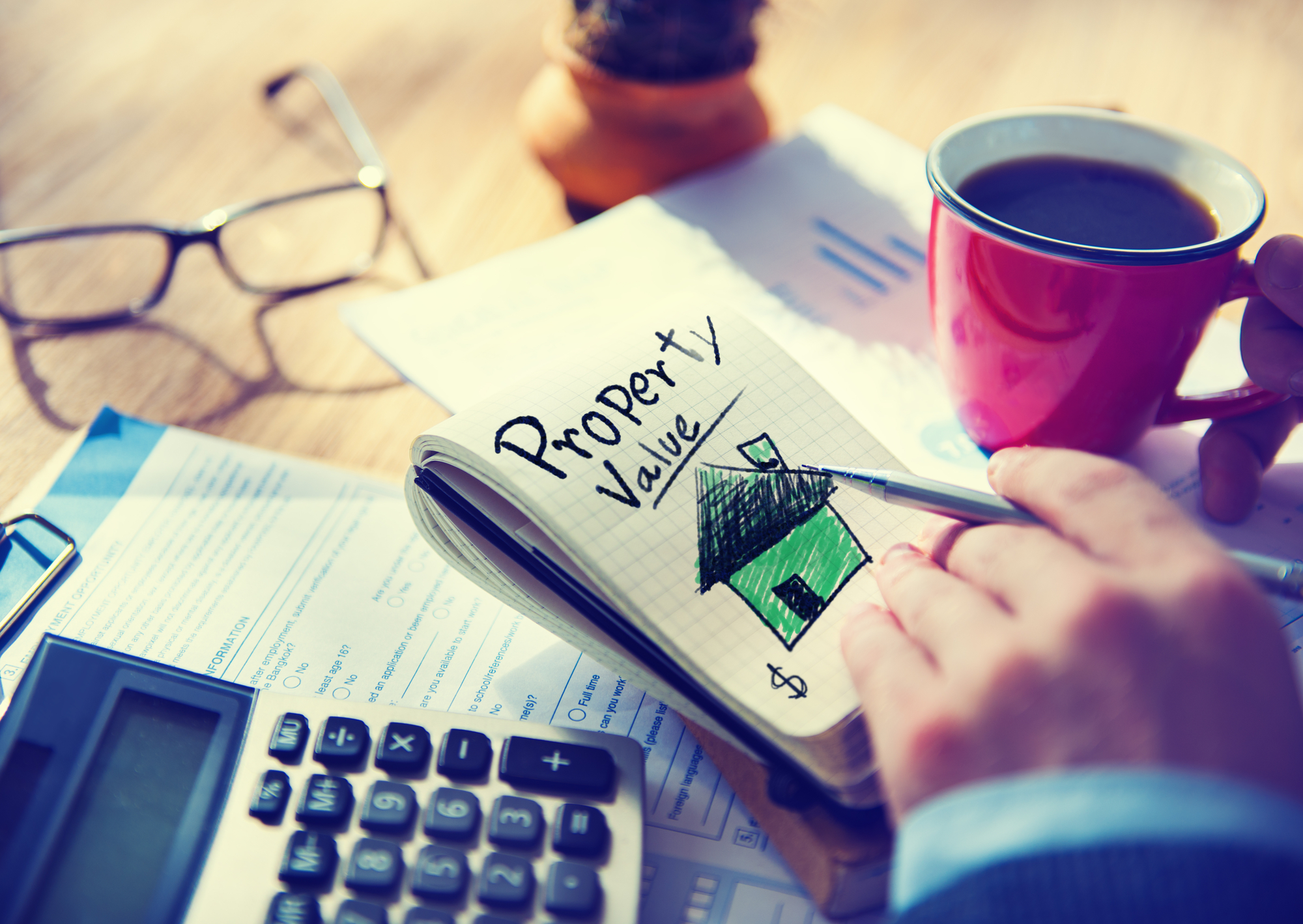 An investor holds a note pad that reads “property value” on their work desk, next to a calculator and financial graphs.