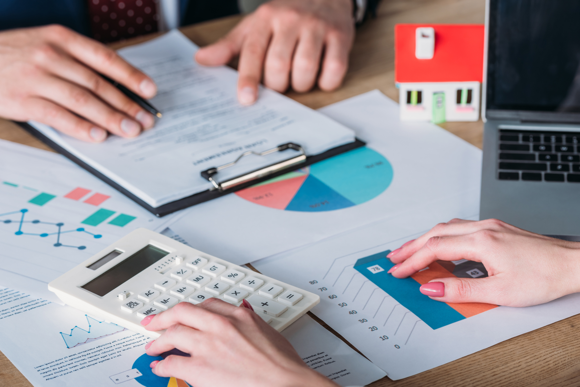 Two people sitting opposite of each other at a desk examining financial graphs with a tiny model house in the background.